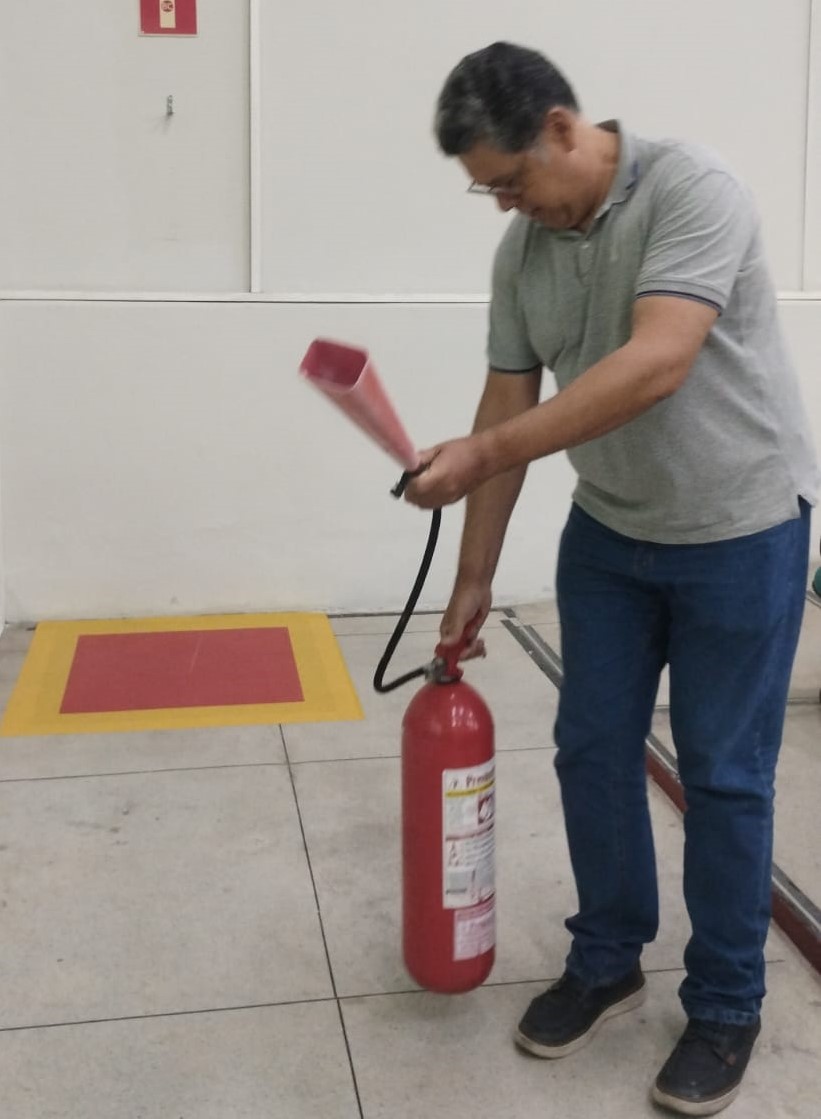 Homem branco, de pé, segurando um hidrante vermelho. O homem usa óculos de grau, veste uma camiseta polo cinza e uma calça jeans. Nos pés, tênis preto.
