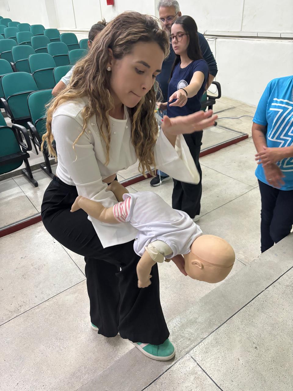 Mulher branca, loira, faz uma manobra de desengasgo em um boneco que simula um bebê. O boneco está de bruços, sobre o braço direito da mulher, o qual está apoiado na sua cocha direita. Com a outra mão espalmada, a mulher bate nas costas do boneco, realizando a manobra de Heimlich. A mulher veste blusa branca e calça preta.