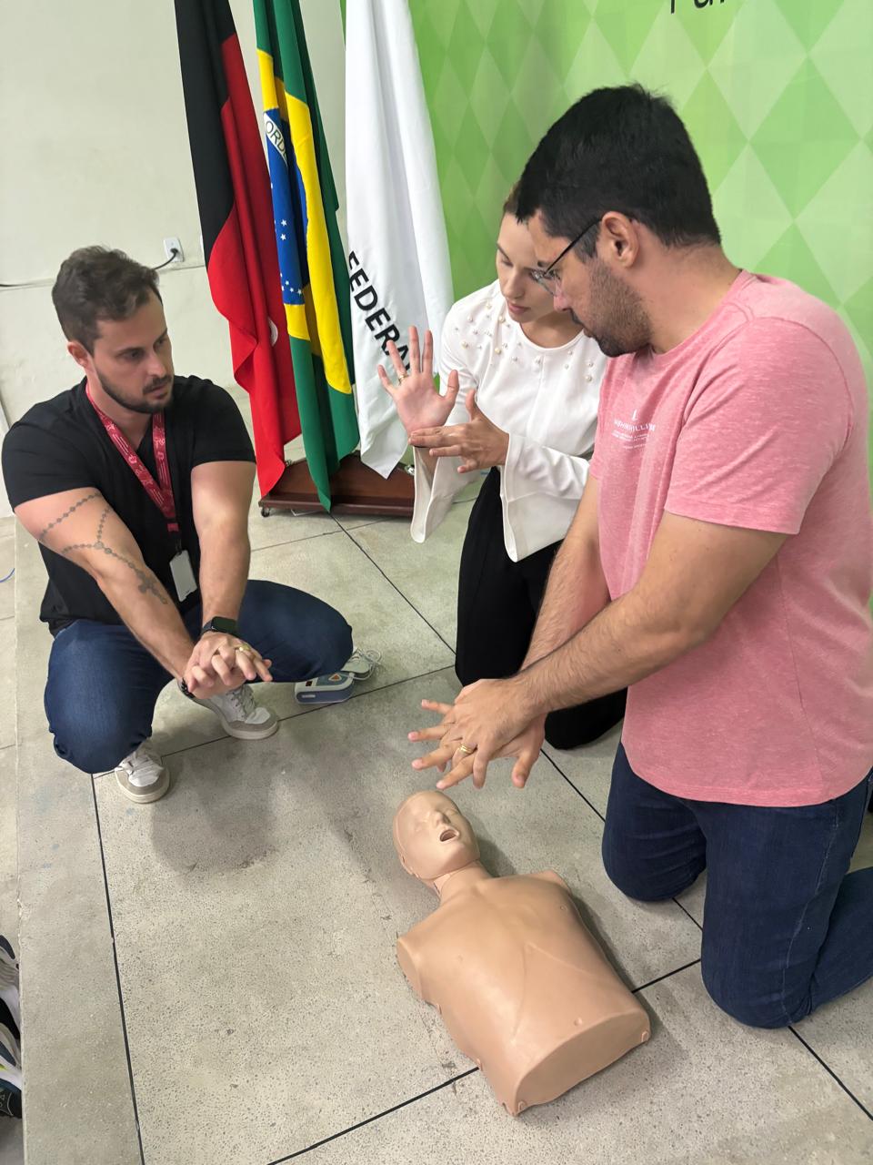 Servidor de joelhos e com as mãos espalmadas, uma sobre a outra, apreende com o instrutor a posição correta de fazer a reanimação cardiopulmonar. No chão, à sua frente, há um boneco de borracha utilizado para o treinamento dessa manobra. Ao lado do servidor, podem ser vistos mais uma servidora e o instrutor, todos de joelhos.